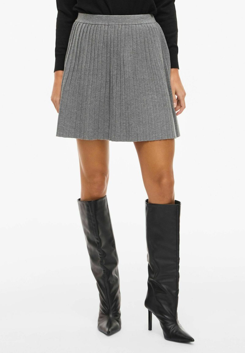 Šedá plisovaná sukně s přiléhavým pasem, vyrobená z měkké látky, doplněná černými kozačkami na podpatku po kolena se špičatou špičkou.