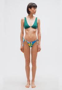 Λεπτή γυναίκα όρθια ξυπόλητη, φοράει πράσινο μπικίνι τοπ και πολύχρωμο φλοράλ κατώτερο μέρος μπικίνι, κοιτάζοντας προς τα αριστερά της σε απλό λευκό φόντο.