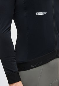 Κοντινό πλάνο μαύρου ποδηλατικού τζέρσεϊ με λογότυπο "SRK Sportful Riders Kit", που δείχνει μακρυμάνικο και ελαστική μέση πάνω από γκρι ποδηλατικά σορτς.