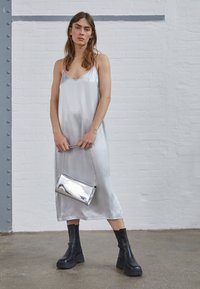 Vestido slip de cetim prateado com decote em V, combinado com uma clutch metálica prateada com alça de corrente e botas de tornozelo pretas com plataforma.