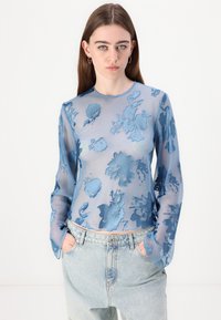 Prosojna plava majica z dolgimi rokavi, okrašena s cvetličnim vzorcem v različnih odtenkih modre, z okroglo ovratnikom in ohlapnim krojem, v kombinaciji s svetlimi denim hlačami.