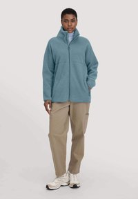 Giacca in pile azzurro chiaro con zip e colletto alto, texture liscia e due tasche laterali, indossata sopra un dolcevita grigio chiaro e pantaloni beige.