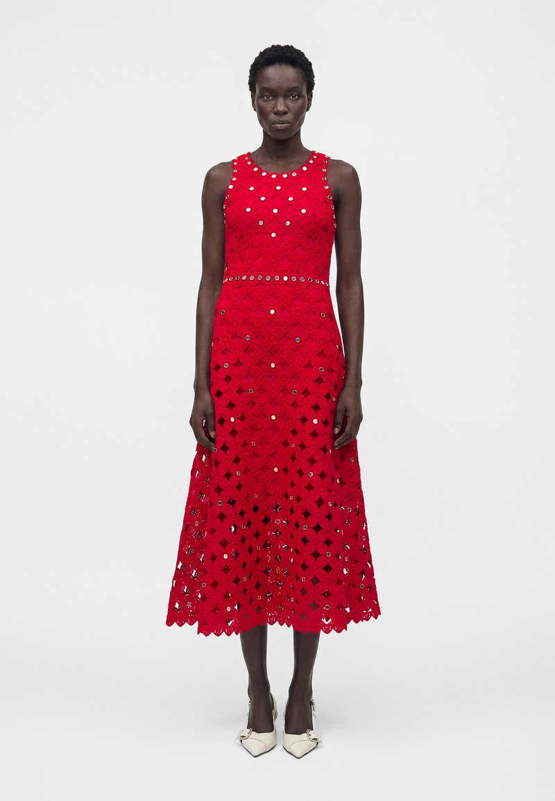 Γυναίκα φοράει αμάνικο κόκκινο μίντι φόρεμα πλεκτό με ασημένιες τρουκς και λευκές ψηλοτάκουνες μυτερές γόβες, στέκεται σε απλό λευκό φόντο.