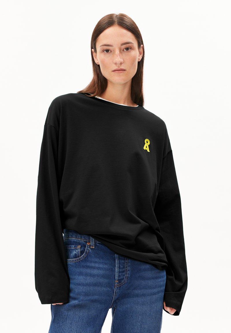 Schwarzes Langarm-T-Shirt mit lockerer Passform, versehen mit einem kleinen gelben Band-Emblem auf der linken Brustseite. Kombiniert mit blauen Jeans.