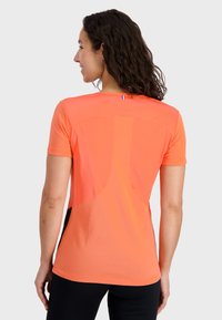 Camiseta atlética de manga corta en naranja brillante, con un diseño texturizado en la parte trasera, un pequeño detalle de bandera en el cuello y paneles laterales en negro.