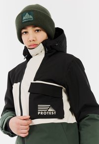 Vodootporna jakna u crnoj, bijeloj i zelenoj boji s visokim ovratnikom, zatvaračem sprijeda i džepom na prsima s logom "Protest". Pleteni zeleni baret.