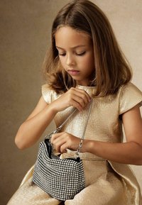 Črna torba okrašena z rhinestones in srebrnim verižnim ročajem, ki jo drži otrok v teksturirani zlati obleki, s poudarkom na podrobnostih torbe.