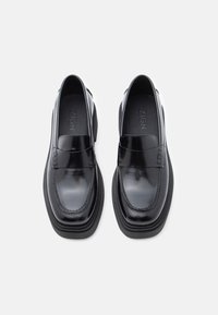 Crni lakirani mokasini s okruglim vrhom, sa šivanim detaljima i debelim crnim gumenenim đonom. Brend "ZIGN" je utisnut unutra.