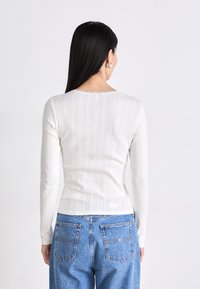 Hvid langærmet top med lodret ribbet tekstur og bølget halsudskæring, parret med højtaljede blå denim jeans med baglommer.