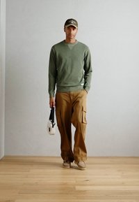 Olijfgroen sweatshirt met lange mouwen, tan cargobroek, beige schoenen en een witte schoudertas. Eenvoudig ontwerp met een ribgebreide ronde hals en een casual pasvorm.