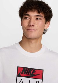 Bela bombažna majica s bold rdečim in črnim Nike logotipom na sredini, okroglim ovratnikom in kratkimi rokavi.