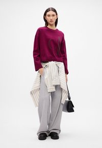 Burgundijas lielizmēra kapuce ar melnu logo, kopā ar pelēkajiem platajiem sporta biksēm. Strīpota krekla josta ap vidukli un melna rokassoma.