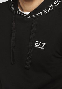 Svart hoodie med en strukturerad design, som har en logotyp på bröstet och en huva med dragsko och mönstrad kant.