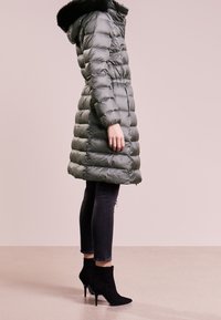 Šedý prošívaný kabát s umělým kožíškem na kapuci, bočními kapsami, vodorovným prošitím a zeštíhleným pasem, doplněný černými kotníkovými botami a těsnými džínami.