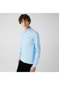 Lys blå langærmet polo-skjorte med krave og et lille grønt logo. Stoffet virker glat og har et klassisk fit design.