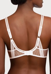 Reggiseno bianco in pizzo con spalline regolabili, decorato con pannelli in rete trasparenti, cuciture delicate, chiusura a gancio e accenti decorativi.