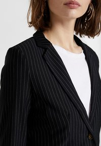 Černý blazer s jemným pruhováním a přiléhavým střihem, vyrobený z hladké tkaniny. Má špičatý límec a zapínání na jeden knoflík. Nositý přes bílou košili.