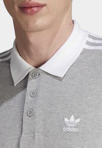 Graues Polohemd mit weißem Kragen, ausgestattet mit drei grauen Knöpfen und einem gestickten weißen Logo auf der Brust. Strukturiertes Material, sportliches Design.