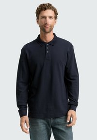 Marineblaues Langarm-Polohemd mit Kragen, einer Drei-Knopf-Leiste und gerippter Textur. Kombiniert mit hellblauen Jeans.