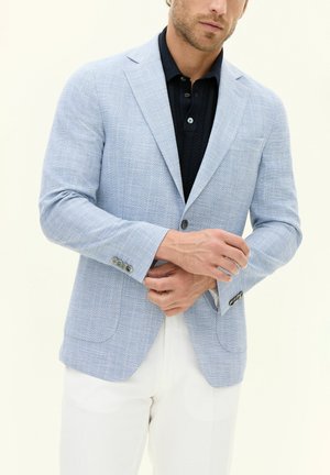 Hellblauer Fischgrätenblazer mit Reverskragen, zwei Knöpfen und subtiler Sheen, über einem dunklen Poloshirt getragen, kombiniert mit weißen Hosen.