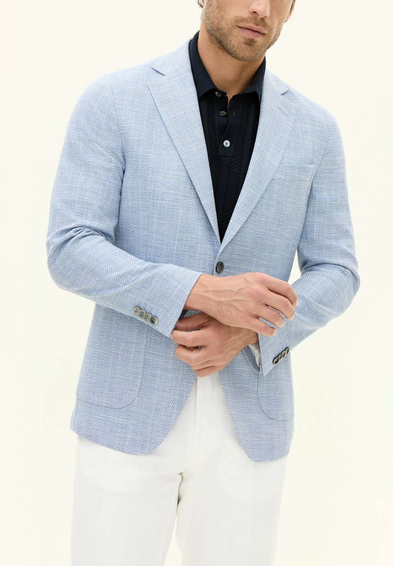 Blazer a rombi azzurro chiaro con rever a punta, due bottoni e una lieve lucentezza, indossato sopra un polo scuro, abbinato a pantaloni bianchi.