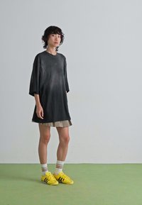 Young person with short curly hair wearing an oversized black shirt, beige skirt, white socks, and bright yellow sneakers standing on a green floor.