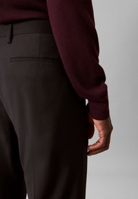 Die Hand eines Mannes ruht neben schwarzen, maßgeschneiderten Hosen mit einem bordeauxfarbenen Langarm-Pullover vor einem schlichten grauen Hintergrund.