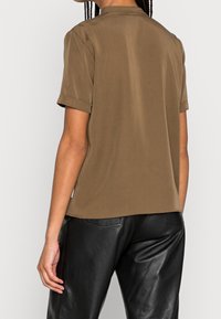 Top marrone a maniche corte con scollatura rotonda, texture liscia e polsini arrotolati, abbinato a pantaloni in pelle nera.