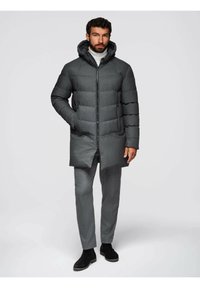 Cappotto imbottito grigio scuro con cappuccio, dotato di zip frontale e tasche laterali, indossato sopra pantaloni grigio chiaro e stivaletti neri alla caviglia.