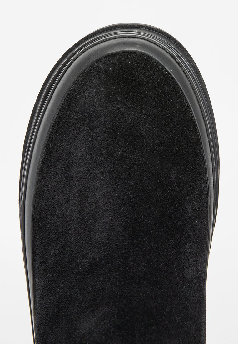 Zapato de ante negro con puntera redondeada, suela negra lisa de contraste, diseño minimalista y contornos limpios. La textura es suave y mate.