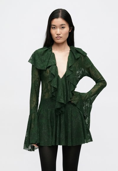 Πράσινο διαφανές φόρεμα με βαθύ V λαιμόκοψη, στρωμένο γιακά με βολάν και μανίκια, με floral μοτίβο και φαρδιές μανσέτες, σε συνδυασμό με σκουρόχρωμο κολάν.