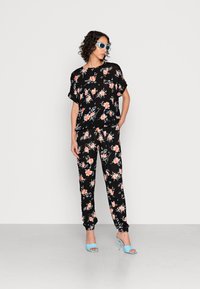 Conjunto de duas peças com padrão floral preto, apresentando uma blusa solta e calças ajustadas. Padrões de rosas brilhantes, tecido suave, combinado com sandálias de salto azuis.