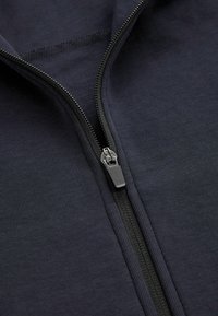 Giacca zip-up blu scuro con una texture liscia, dotata di una cerniera metallica e di una fodera interna visibile attraverso la zip aperta.