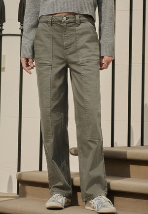 Persona ģērbta zaļās taisnās biksēs ar priekšējiem ielāpiem, pelēkā īsā džemperī un pelēkās sporta kurpēs, stāv uz kāpnēm.