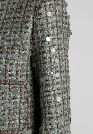 Giacca in tweed testurizzato verde menta con accenti rossi, caratterizzata da paillettes metalliche rotonde cucite nel tessuto. Una tasca sul petto visibile.