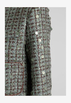 Giacca in tweed testurizzato verde menta con accenti rossi, caratterizzata da paillettes metalliche rotonde cucite nel tessuto. Una tasca sul petto visibile.