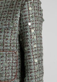 Texturierte Tweedjacke in Mintgrün mit roten Akzenten, die runde, metallicfarbene Pailletten enthält, die in den Stoff genäht sind. Eine Brusttasche sichtbar.
