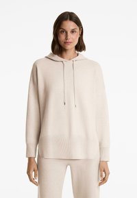 Femme portant un sweat à capuche beige oversize avec des cordons et un pantalon beige assorti, se tenant devant un fond blanc.