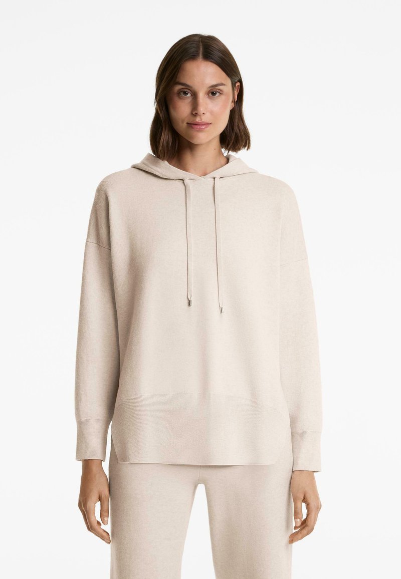 Femme portant un sweat à capuche beige oversize avec des cordons et un pantalon beige assorti, se tenant devant un fond blanc.