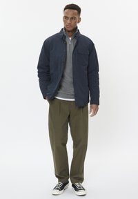 Chaqueta externa navy con un bolsillo delantero, superpuesta a una sudadera gris con cremallera, combinada con pantalones verdes oliva y zapatillas negras.