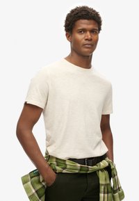 Crème-kleurige T-shirt met korte mouwen en een ronde hals, gecombineerd met een groene en geruite blouse die om de taille is geknoopt, gedragen met donkere broek.