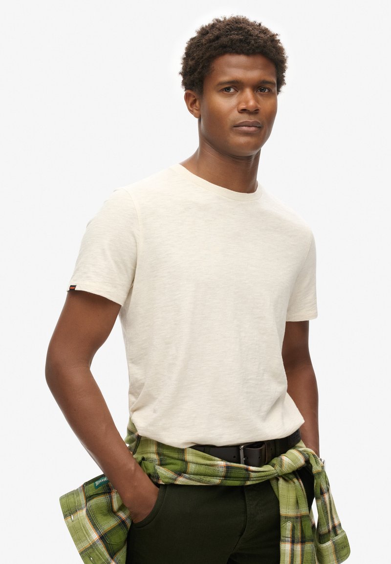 Crème-kleurige T-shirt met korte mouwen en een ronde hals, gecombineerd met een groene en geruite blouse die om de taille is geknoopt, gedragen met donkere broek.