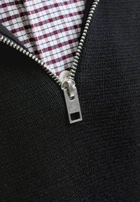 Fermeture éclair argentée partiellement ouverte sur un vêtement en maille noir, révélant une chemise blanche à motif à carreaux rouges et bleus en dessous.