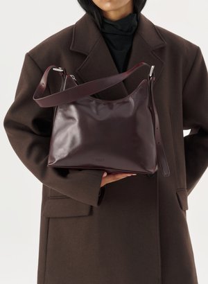 Person in dark brown coat holding a glossy dark burgundy leather handbag with adjustable shoulder strap against a light background.