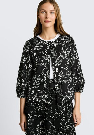 Junge Frau trägt schwarze und weiße, blumige, knöpfbare Jacke mit Dreiviertelärmeln und passenden gemusterten Rock, steht vor einfarbigem Hintergrund.