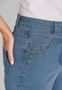 Blaue Denim-Jeans mit mittlerer Taille, verziert mit goldenen Nähten, einer seitlichen Tasche mit Klappe und einer Hand, die auf der Hüfte ruht.