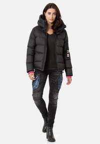 Schwarze Pufferjacke mit Kapuze, ausgestattet mit horizontalen Steppnähten, rosa Akzenten und einem Logo. Kombiniert mit ausgewaschenen schwarzen Jeans und schwarzen Stiefeln.