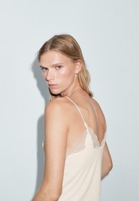 Camisole beige clair avec une délicate bordure en dentelle le long de l'encolure. Dotée de fines bretelles ajustables et d'une texture douce et lisse. Vue de dos présentée.