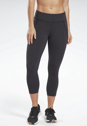 Personne au ventre nu portant des leggings noirs courts et des chaussures de sport noires, debout devant un fond blanc.