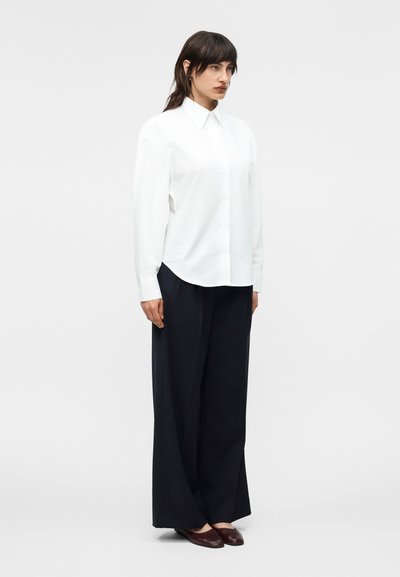 Γυναίκα που στέκεται φορώντας μια φαρδιά λευκή μακρυμάνικη πουκάμισο, φαρδιά μαύρη παντελόνια και καφέ επίπεδα παπούτσια μπροστά σε έναν απλό ανοιχτό φόντο.